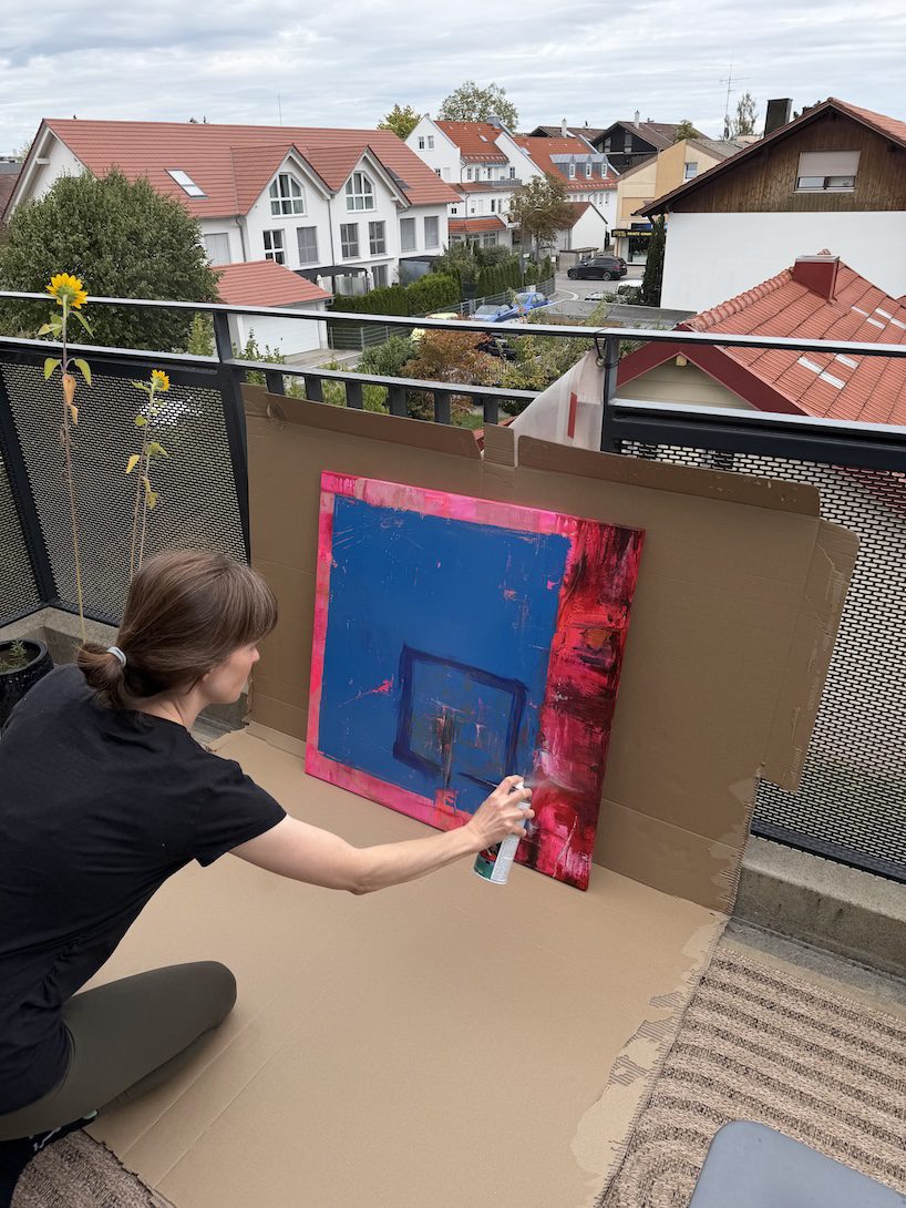 Künstlerin Anika Adam beim Auftragen von Firnis auf ein großformatiges, abstraktes Acrylgemälde in Blau und Pink auf dem Atelier-Balkon